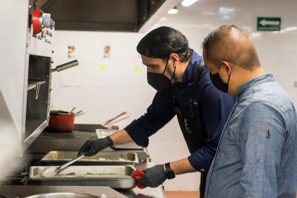 Two chefs in masks and gloves cooking in a professional kitchen setting.